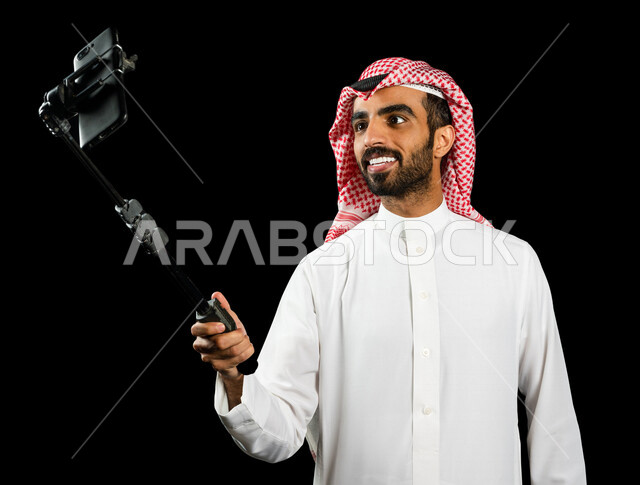 رجل عربي خليجي سعودي يرتدي الزي السعودي يقوم بالتقاط صور سيلفي من خلال الهاتف المحمول، استخدام حامل الجوال لتصوير سلفي, أخذ صوره شخصيه, صور تذكاريه, لحظات و ذكريات جميله, خلفيه سوداء