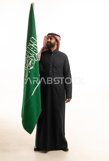 رجل سعودي بالثوب التقليدي يقف مع علم السعودية