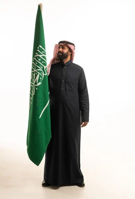 رجل سعودي بالثوب التقليدي يقف مع علم السعودية