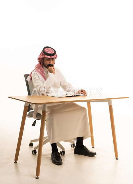 رجل سعودي بالزي التقليدي يقرأ كتاباً خلف المكتب