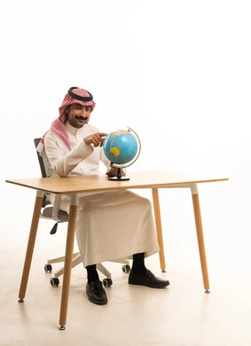 Saudi Man in Traditional Thobe Pointing at Globe at Desk