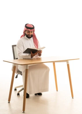 رجل سعودي بالزي التقليدي يقرأ كتاباً خلف المكتب