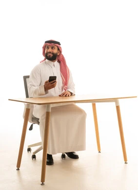 رجل أعمال سعودي ممسك بهاتف ذكي خلف مكتبه