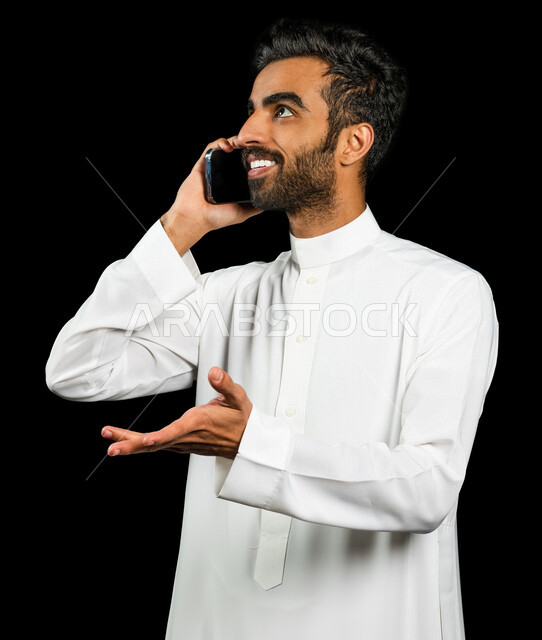 A Saudi Arabian Gulf man makes a call through a mobile phone, make a phone call, contact and acquaintance, chat and conversations, social relations, facial gestures indicating happiness, black background