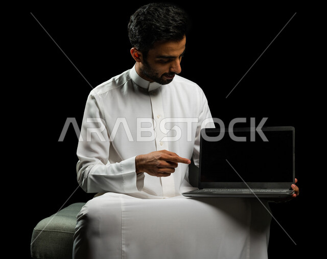 رجل عربي سعودي خليجي يرتدي الزي السعودي، إستخدام جهاز اللابتوب لإنجاز المهام، التسوق الإلكتروني، تطور التكنولوجيا وإستخدام الأجهزة التقنية الحديثة، إجتماع عمل، إدارة الأعمال عن بعد، تصفح الانترنت, إجراء مكالمات فيديو عبر جهاز الحاسوب, خلفيه سوداء