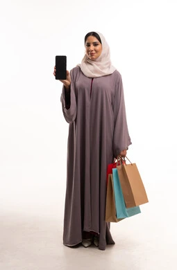 امرأة سعودية تحمل أكياس تسوق وهاتف ذكي بخلفية بيضاء