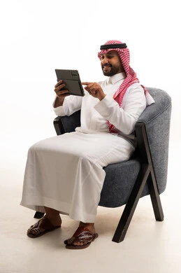 رجل سعودي يستخدم جهازاً لوحياً على كرسي بخلفية بيضاء