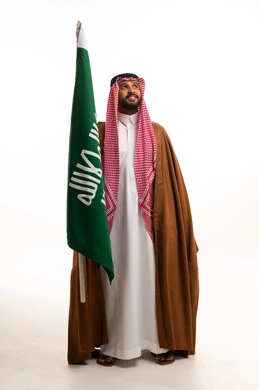 رجل سعودي بالزي التقليدي يحمل علم السعودية بخلفية بيضاء