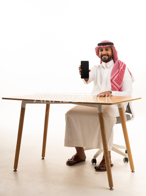رجل سعودي بالثوب التقليدي يمسك هاتف ذكي على مكتب