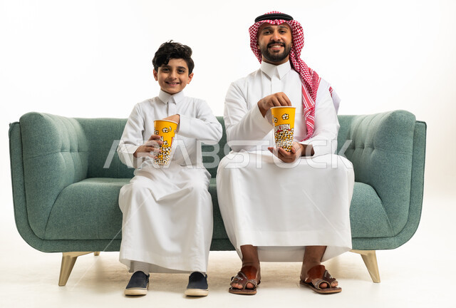أب سعودي وابنه يتناولان الفشار على أريكة خضراء في استوديو