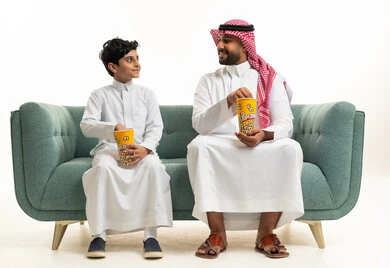 رجل سعودي وطفل يتناولان الفشار على أريكة خضراء
