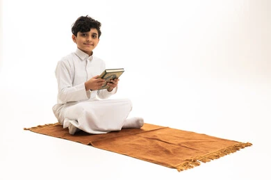 طفل سعودي بالثوب الأبيض يحمل المصحف على سجادة الصلاة