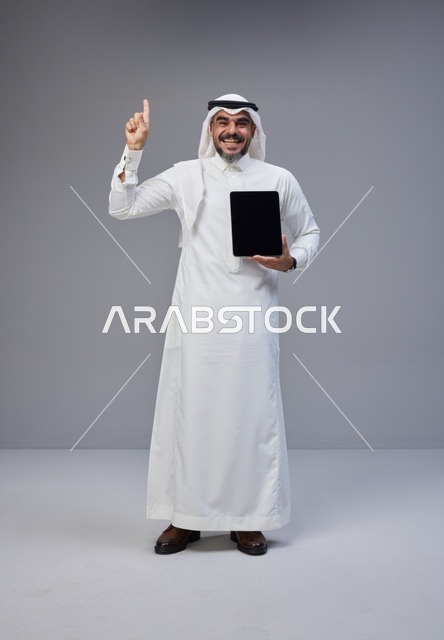 Saudi Man Holding Tablet Pointing Up on Grey Background