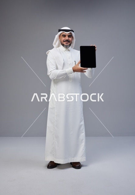 رجل سعودي بالزي التقليدي يحمل جهاز لوحي بشاشة فارغة