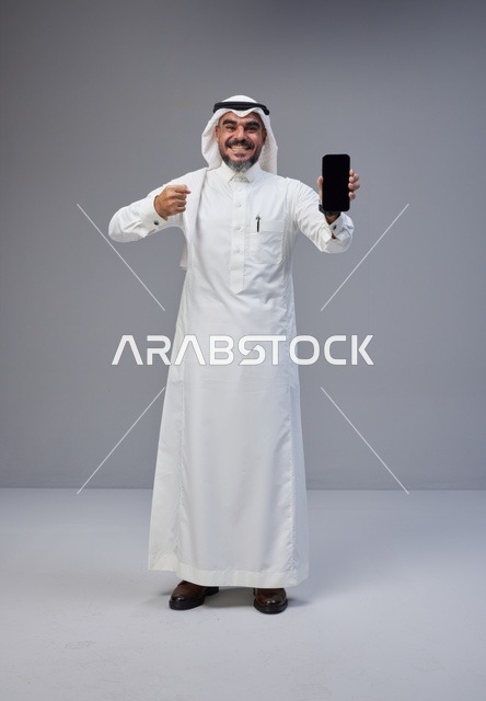 رجل سعودي بالزي التقليدي يحمل هاتفاً ذكياً بشاشة فارغة