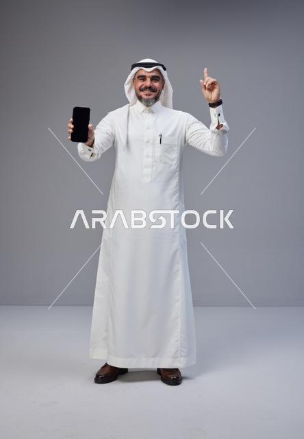 رجل سعودي يحمل هاتفاً بشاشة فارغة على خلفية رمادية