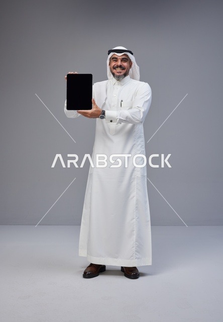 رجل سعودي بالزي التقليدي يحمل جهاز لوحي بشاشة فارغة