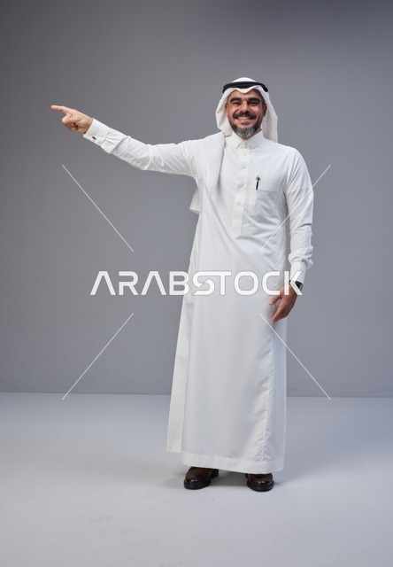 رجل سعودي بالثوب التقليدي يشير بيده في استوديو خلفية رمادية
