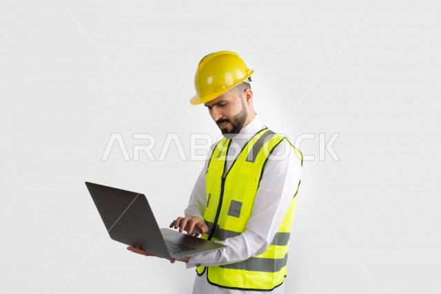 مهندس معماري عربي سعودي  ، استخدام الكمبيوتر المحمول ، رجل  انشاءات باللباس السعودي ، خوذة المهندس ،خلفية بيضاء