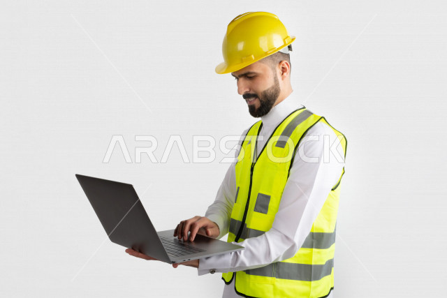 مهندس معماري عربي سعودي  ، استخدام الكمبيوتر المحمول ، رجل  انشاءات باللباس السعودي ، خوذة المهندس ،خلفية بيضاء