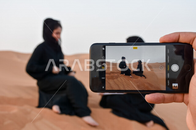 صورة مقربة ليد شخص يمسك هاتف محمول ويلتقط صورة لفتاتين عربيتان خليجيتان ترتديان عباية معاصرة في صحراء العين بأبو ظبي، الكثبان الرملية، السياحة في الإمارات العربية المتحدة