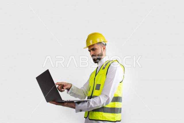 مهندس معماري عربي سعودي  ، استخدام الكمبيوتر المحمول ، رجل  انشاءات باللباس السعودي ، خوذة المهندس ،خلفية بيضاء