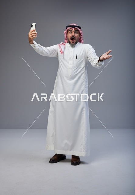 رجل سعودي بالزي التقليدي مندهش يمسك هاتف ذكي