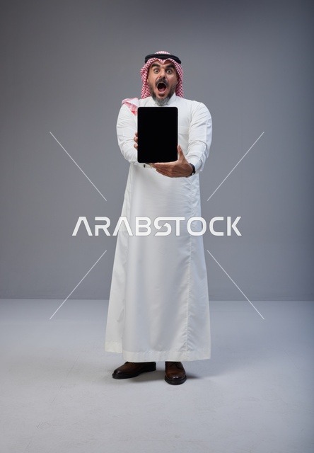 رجل سعودي يحمل جهاز لوحي بتعبير وجه مصدوم في الاستوديو