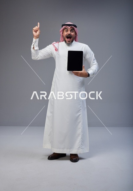 رجل سعودي يحمل جهاز لوحي ويشير للأعلى بخلفية رمادية