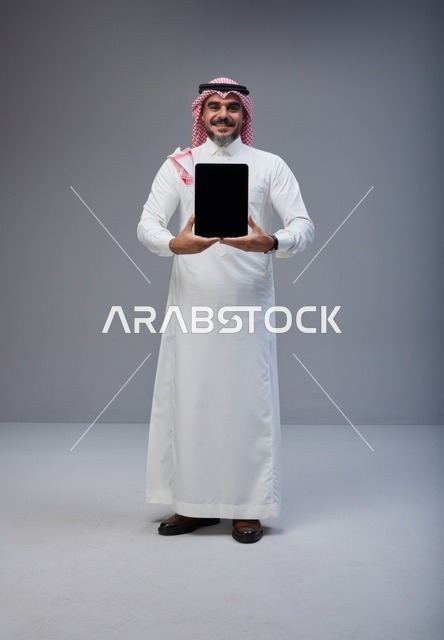 رجل سعودي بالزي التقليدي يحمل جهاز لوحي بشاشة فارغة