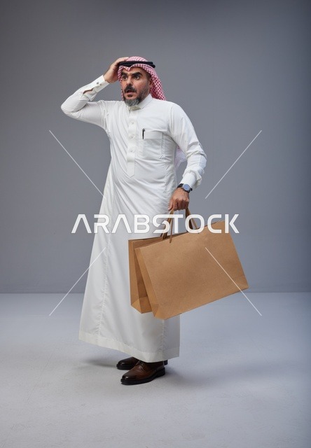 رجل سعودي بالزي التقليدي مصدوم يحمل أكياس تسوق ورقية