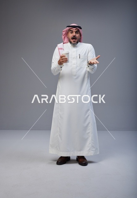 رجل سعودي مندهش يحمل كوب قهوة في استوديو رمادي