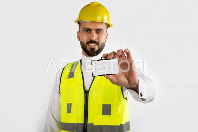 مهندس معماري سعودي مبتسم بيده بطاقة تعريفيه بيضاء  ، خوذة عمل صفراء ، خلفية بيضاء