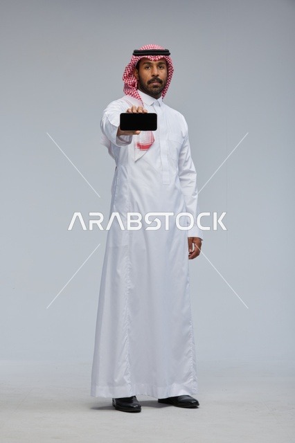رجل سعودي يعرض هاتفاً ذكياً في استوديو بخلفية رمادية