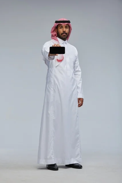 رجل سعودي يعرض هاتفاً ذكياً في استوديو بخلفية رمادية