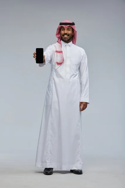 رجل سعودي يحمل هاتف ذكي في استوديو تصوير بخلفية رمادية