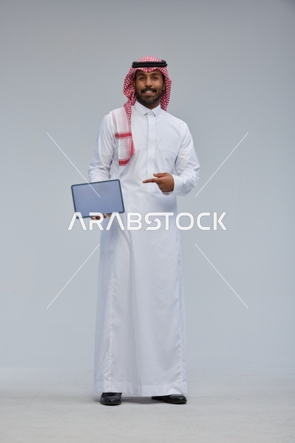رجل سعودي مبتسم يحمل جهازاً لوحياً بالزي التقليدي