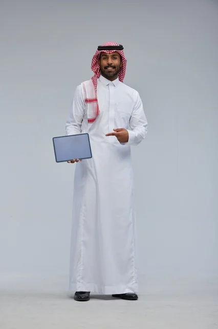 رجل سعودي مبتسم يحمل جهازاً لوحياً بالزي التقليدي