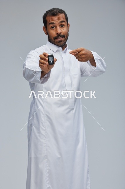 رجل سعودي بالثوب يحمل مفتاح سيارة بخلفية رمادية