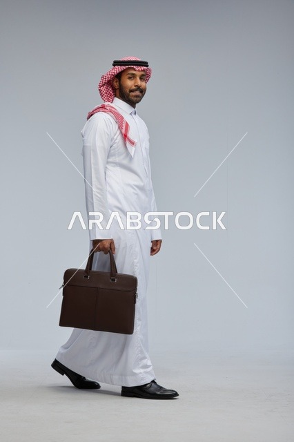 رجل أعمال سعودي يمشي بحقيبة عمل على خلفية رمادية