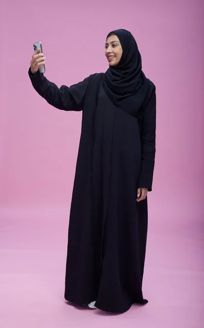 امرأة سعودية بالعباءة تلتقط سيلفي أمام خلفية وردية
