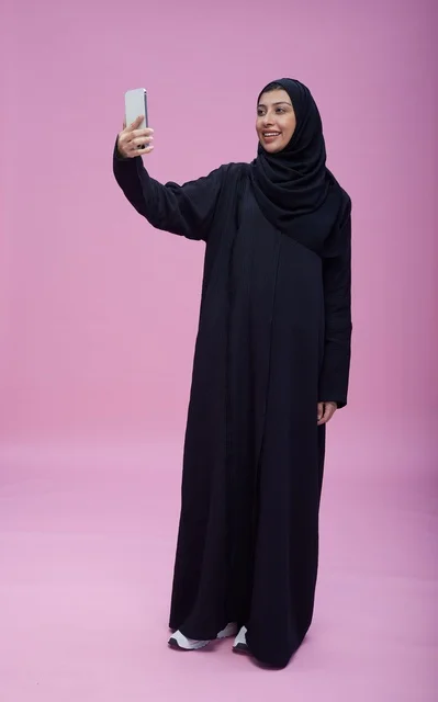 امرأة سعودية بالعباءة تلتقط سيلفي أمام خلفية وردية