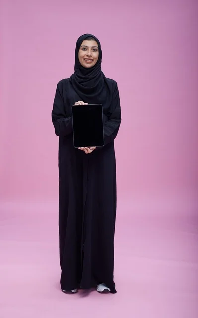 امرأة سعودية تحمل جهازاً لوحياً أمام خلفية وردية