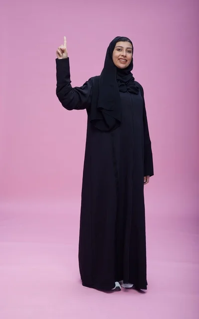 امرأة سعودية بالعباءة تشير للأعلى في استوديو بخلفية وردية