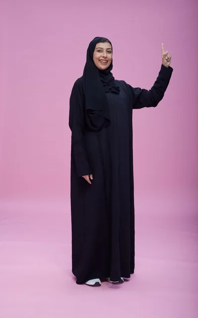 امرأة سعودية بالعباءة تشير للأعلى بخلفية وردية