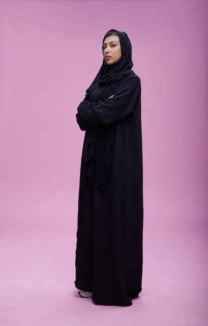 امرأة سعودية بالعباءة تقف بثقة أمام خلفية وردية
