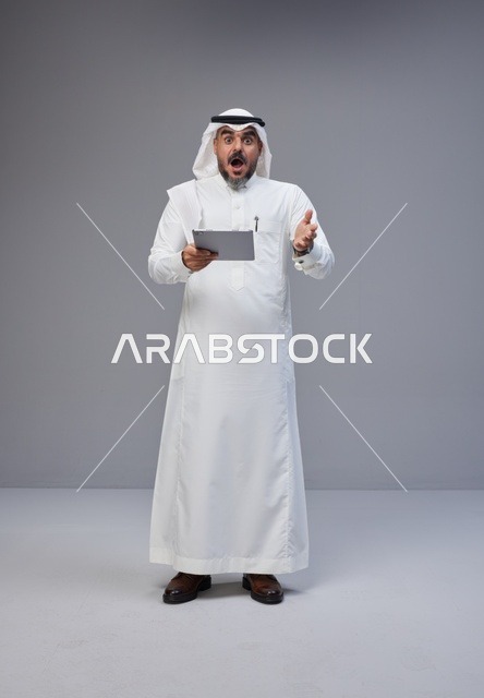 رجل سعودي متفاجئ يحمل جهاز لوحي بخلفية رمادية