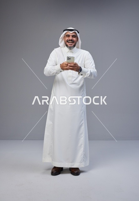 رجل سعودي بالزي التقليدي يضحك ويستخدم الهاتف الذكي