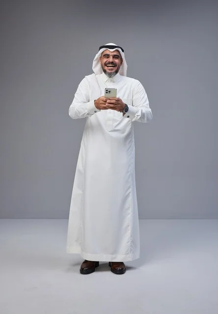 رجل سعودي بالزي التقليدي يضحك ويستخدم الهاتف الذكي
