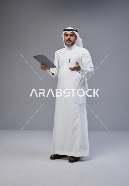 رجل سعودي بالزي التقليدي يمسك جهازاً لوحياً في استوديو